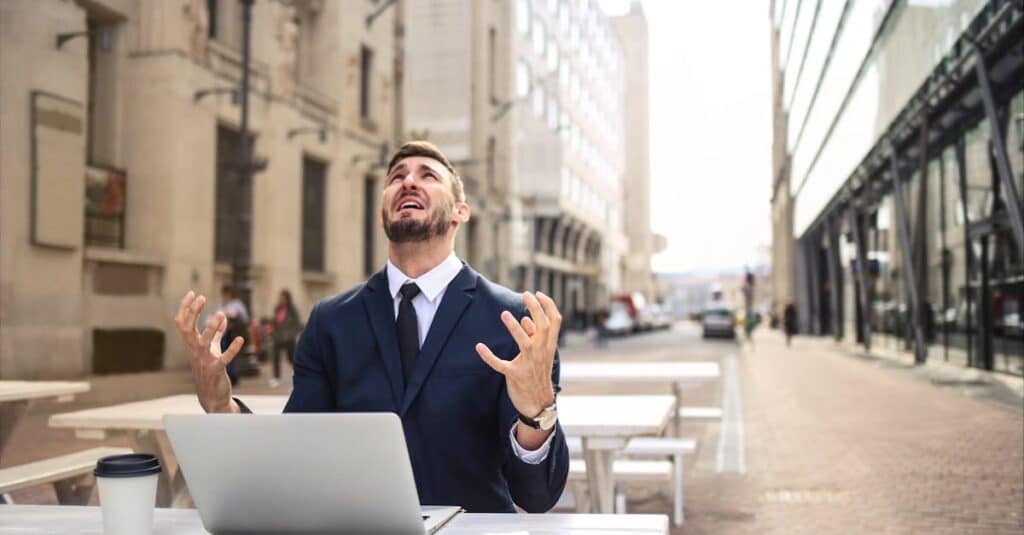 Businessman in suit showing frustration at outdoor table with laptop and coffee.